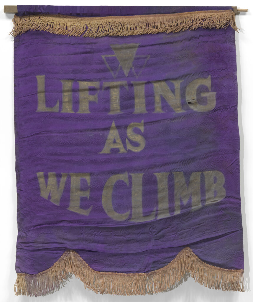 A purple silk banner with gold fringe and the National Association of Colored Women's Clubs' motto, 'LIFTING / AS / WE CLIMB' painted in large gold letters.