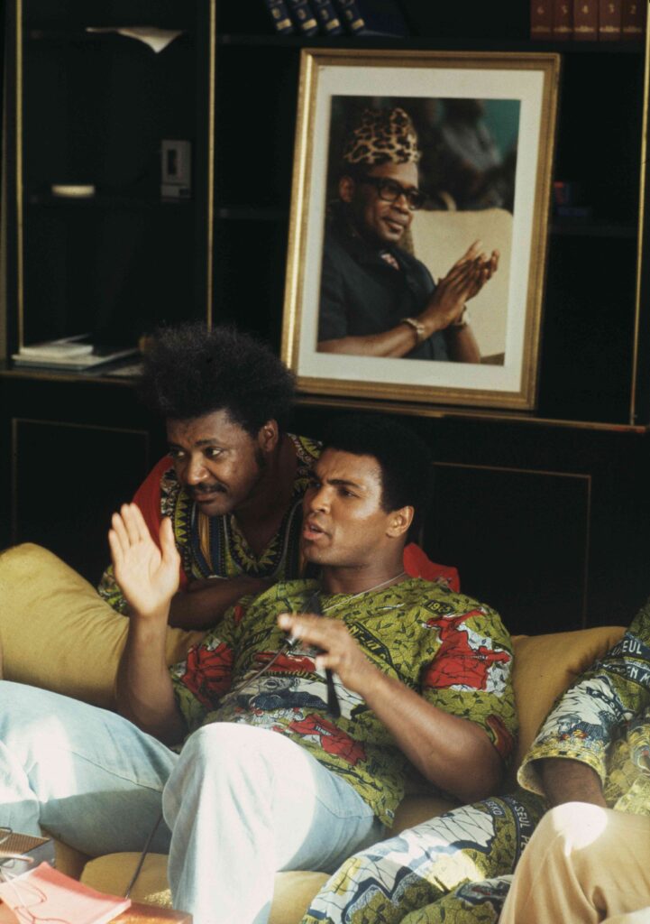 Heavyweight boxer Muhammad Ali (right, 1942 - 2016) and fight promoter Don King relax in Ali's villa ahead of his fight against champion George Foreman in Kinshasa, Zaire, October 26th 1974. On the wall behind them is a portrait of Mobutu Sese Seko, the President of Zaire.