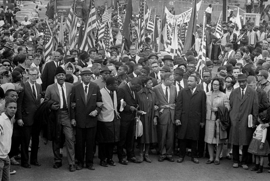 Dr. Martin Luther King (center) leads an estimated 10,00 or more civil rights marchers out on the last leg of their Selma-to-Montgomery march. Others identifiable in front row include: John Lewis, (2d from left) of SNCC; King's aide, Reverend Ralph Abernathy (3rd from left); Dr. Ralph Bunche (5th from left, looking to side); Mrs. King (next to King); and Rev. Hosea Williams (carrying little girl, right).