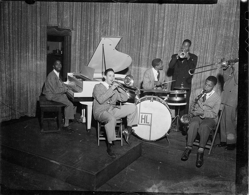 Jazz band performing with Ahmad Jamal on piano, Jon Morris on trombone, Harold 'Brushes' Lee on drums, Horace Turner on trumpet, John Foster on saxophone, and Sam Hurt (cut off on right) on trombone, in stage in front of sparkled curtain, Pittsburgh, Pennsylvania, 1945.